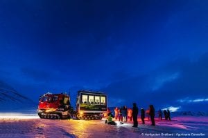 Snowcat with guide and people waiting outide behind it in dark blue winter landscape of Svalbard, Arctic Norway