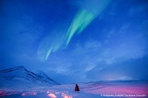 Person sitting on pink snowy ground in distance with back facing camera looking up at light northern lights show in light blue cloudy sky in Svalbard, Arctic Norway