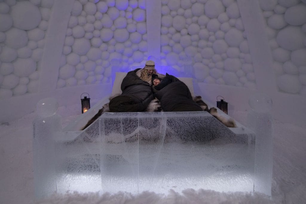 Couple cuddling in sleeping bags on reindeer skins on ice bed in ice hotel room of ice domes in Arctic Norway