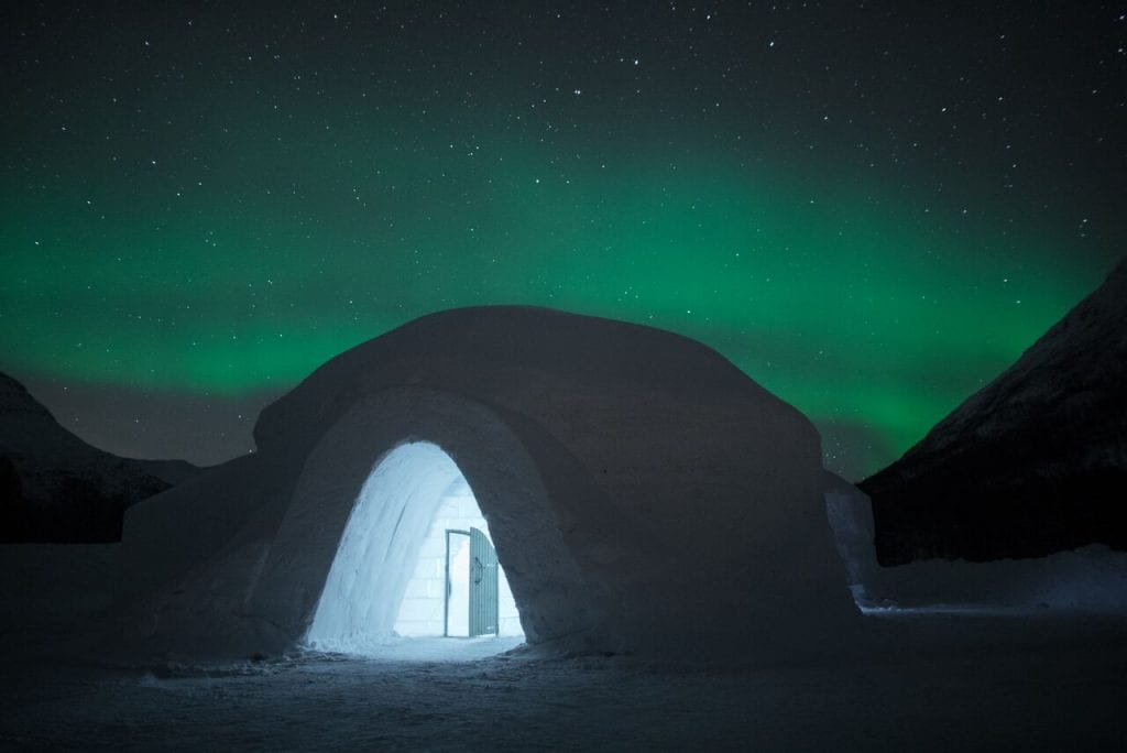 Ice dome exterior with view of lit up entrance and door in front of starry night sky and green northern lights in Arctic Norway