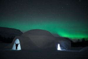 Ice dome exterior with view of lit up entrance and silhouette of person holding arms up in front of starry night sky and green northern lights in Arctic Norway