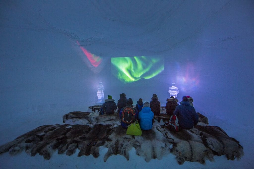 People sitting on reindeer skins in ice room with backs turned watching screening of northern lights video in ice domes of Arctic Norway