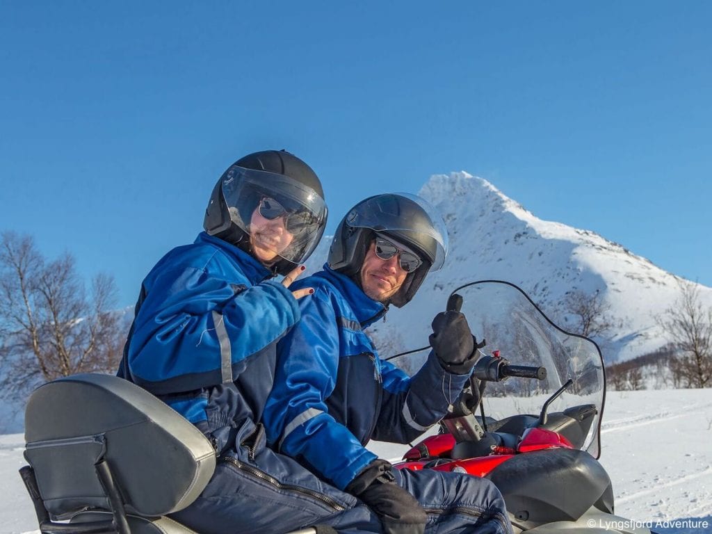 Couple on snowmobile smiling back at camera with shades and helmets giving peace sign and thumbs up in the mountains of Lyngen, Arctic Norway