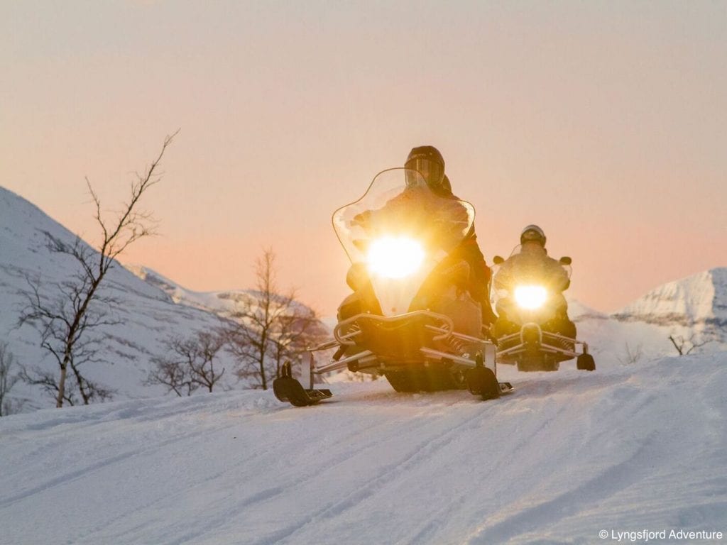 Two snowmobiles with lights on driving towards camera in front of pink winter sky in Lyngen, Arctic Norway