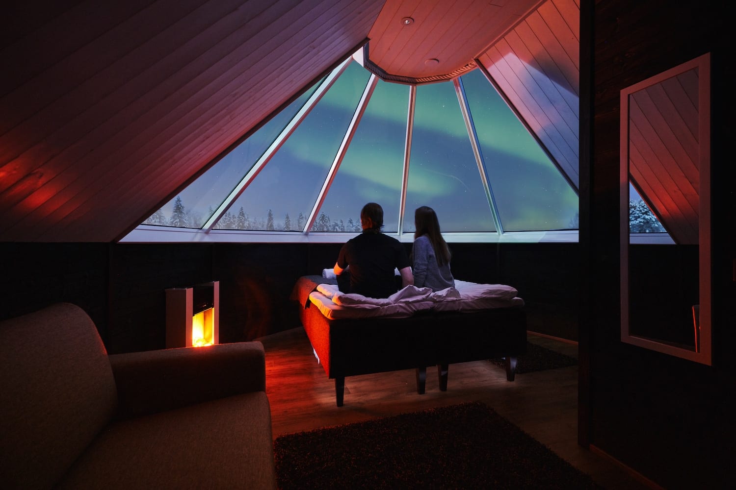 Couple sitting on bed in cosy aurora cabin looking out glass window at northern lights