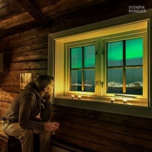 Man sitting in fishermen rorbuer cabin at night looking out of window at northern lights in svolvær, norway