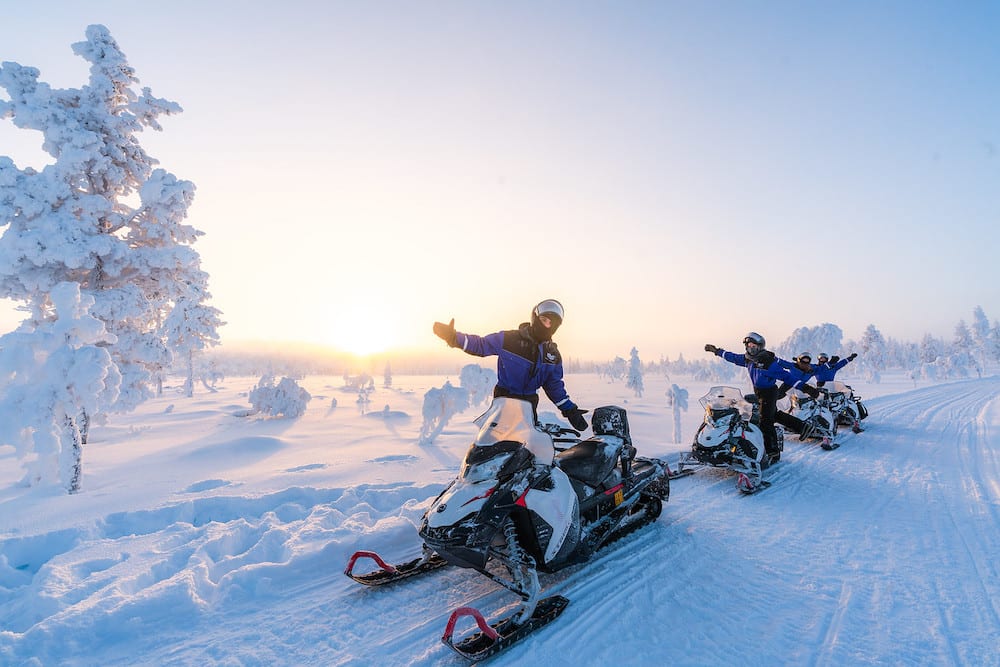 People on top of snowmobiles posing for photograph with snow covered trees and snow on ground with sun shining bright